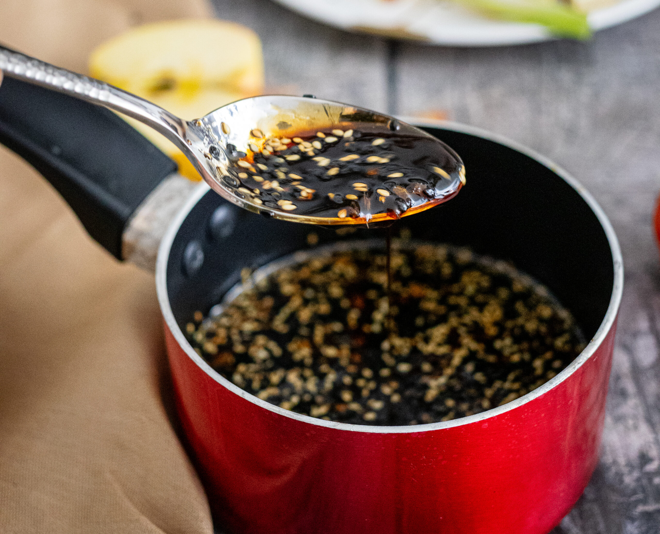 Löffel mit dunkler Soße und Sesam über einem roten Topf, auf Holztisch mit Apfelstück im Hintergrund.