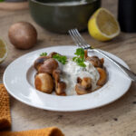 Ein Teller mit gebratenen Pilzen in einer weißen Sahnesauce, garniert mit Petersilie; im Hintergrund liegen Zitronenhälften.