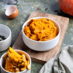 Weiße Schale mit Kürbispüree auf Holzbrett, daneben Kürbisstück mit Kernen, Ei und Kürbiskerne in kleiner Schale.