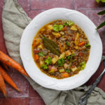 Teller mit Linsensuppe, dekoriert mit Kräutern. Daneben frische Karotten und ein Löffel auf einem Tuch.
