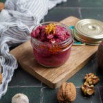 Rotes Pesto in einem Glas auf Holzbrett, umgeben von Walnüssen, Knoblauch, Roter Bete-Hälften und einem gestreiften Stofftuch.