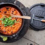 Ein gusseiserner Topf mit rotem Gemüseeintopf, garniert mit Koriander, daneben ein Deckel mit Griff und eine Limette.