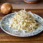 Teller mit Sauerkraut und Dill, daneben eine Zwiebel und eine Gabel auf einem Holztisch.