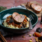 Schüssel mit Couscous, Lammfleisch, Gemüse und orientalischen Gewürzen auf einem Tisch mit Zimtstangen und Trockenfrüchten.