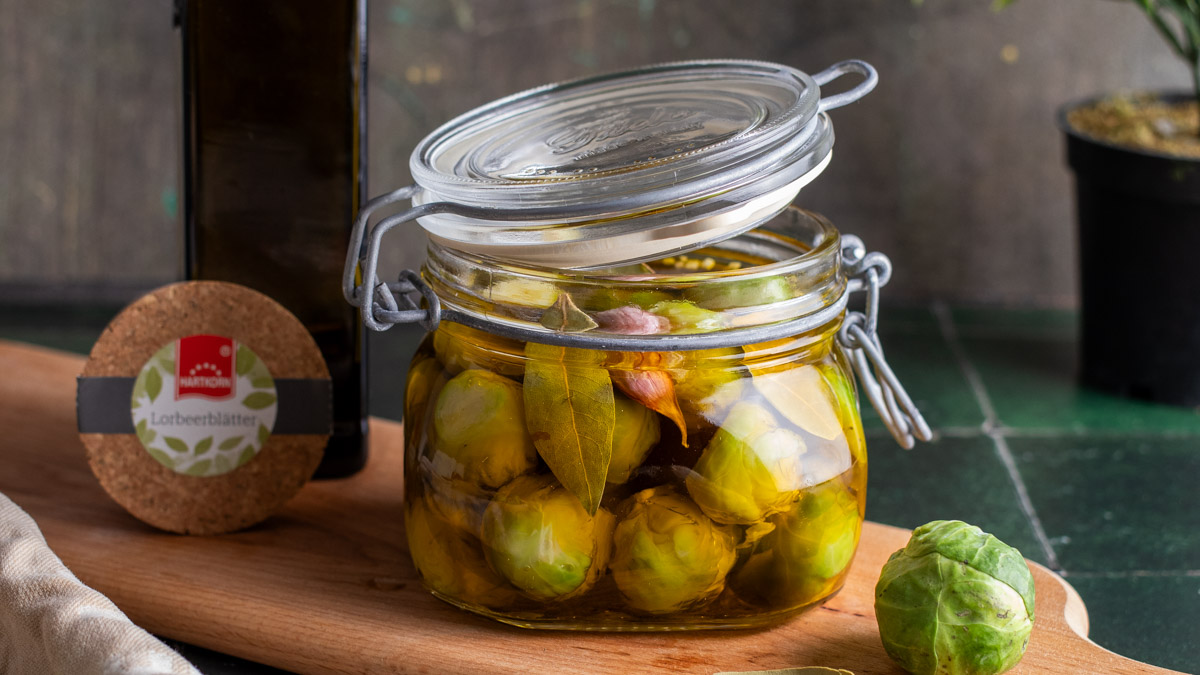 Ein geöffnetes Glas mit eingelegtem Rosenkohl in Öl, daneben Lorbeerblätter und ein frischer Rosenkohl auf einem Holzbrett.
