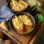 Zwiebelnsuppe mit Käse überbackenem Brot in Schalen auf Holzbrett, daneben Zwiebeln, Bayblätter und geriebener Käse.