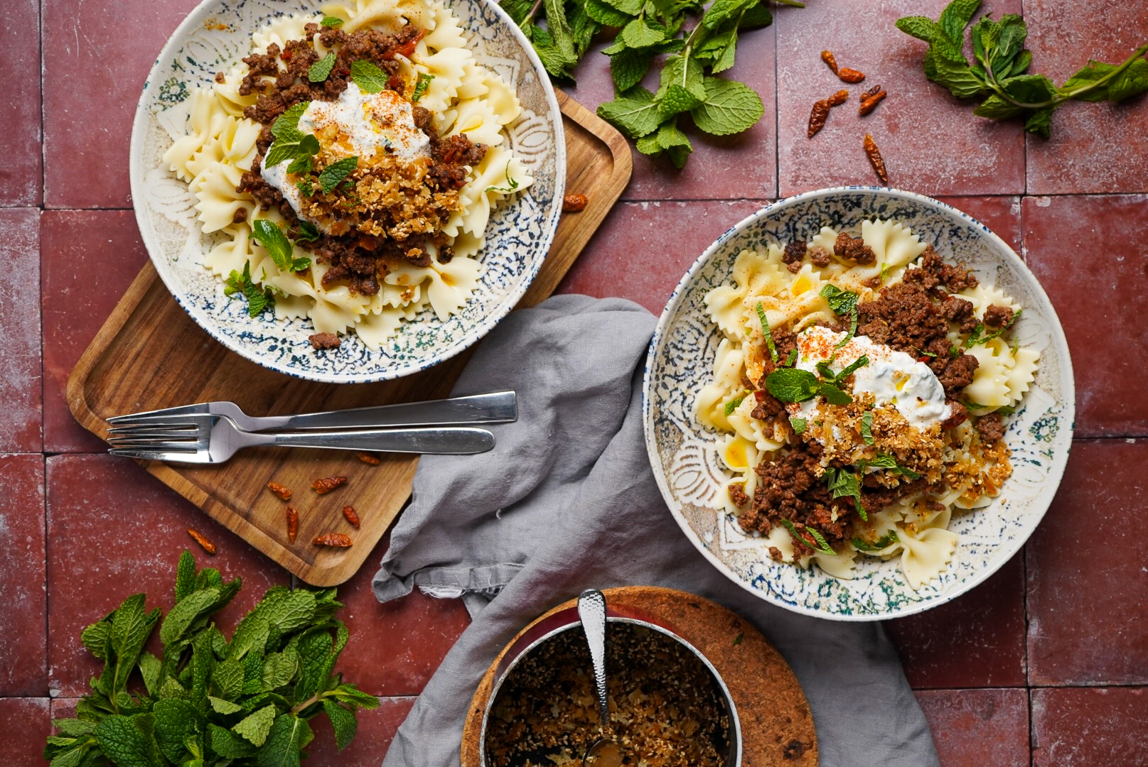 Türkische Pasta 2.0 Zwei Teller mit Farfalle, Hackfleisch und Joghurt, garniert mit Kräutern auf einem Holzbrett, umgeben von Minzblättern.