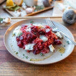 Teller mit Rote-Bete-Risotto und Blauschimmelkäse, garniert mit Kräutern, auf Holzplatte, im Hintergrund Zutaten.
