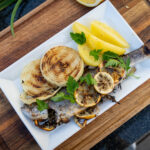 Gegrillter Fisch mit Zitronenscheiben, Zwiebeln und Kartoffelspalten auf weißem Teller, dekoriert mit Petersilie.