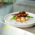 Gericht mit Reis, gegrilltem Fleisch, grünen Bohnen und Erdnüssen auf weißem Teller, dekoriert mit roten Fäden.