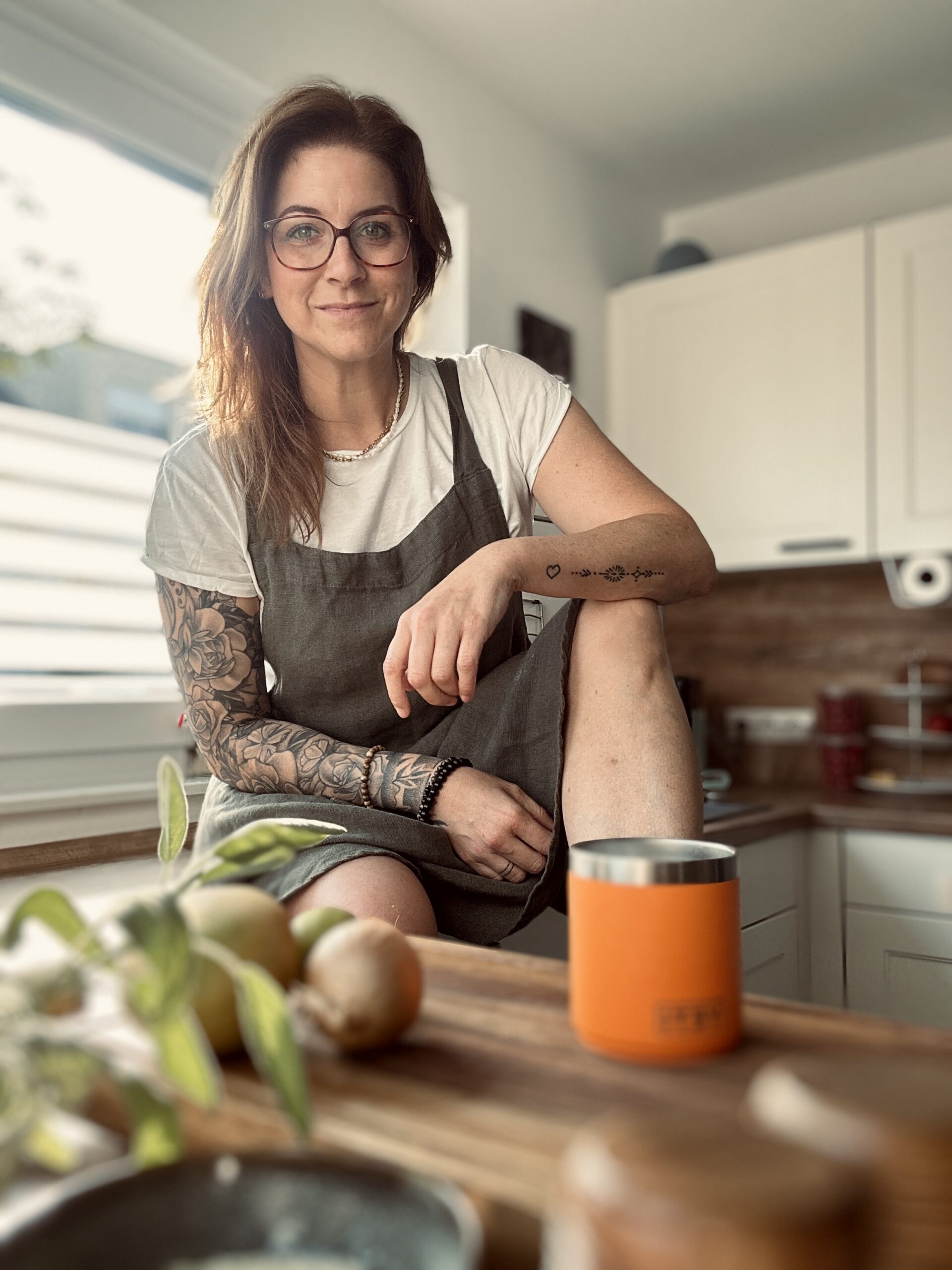 Christina Becher Person mit Brille und Tattoos sitzt in einer Küche auf der Arbeitsplatte, trägt Schürze. Im Vordergrund liegen Zwiebeln.