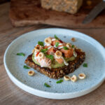 Vollkornbrot mit Avocado, Lachs, Haselnüssen und grünem Blattwerk auf weißem Teller serviert.