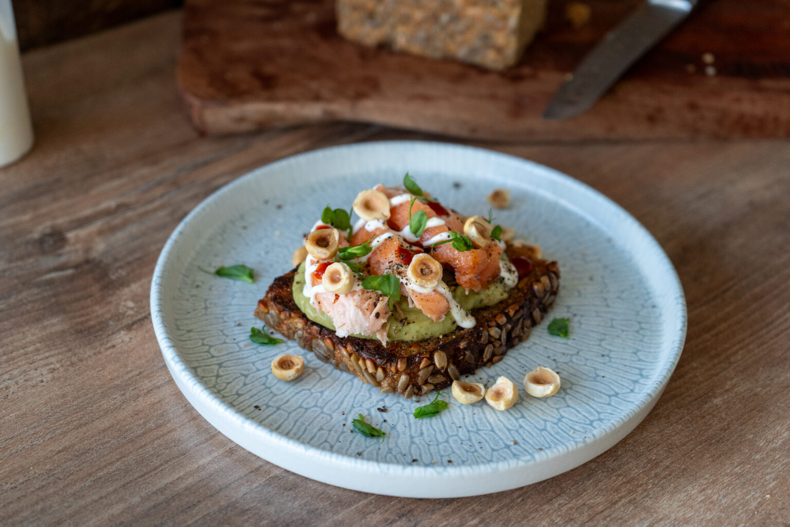 Vollkornbrot mit Avocado, Lachs, Haselnüssen und grünem Blattwerk auf weißem Teller serviert.