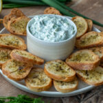 Ein Teller mit gerösteten Brotscheiben und einer Schale Kräuterdip, umgeben von Zitrone, Knoblauch und Kräutern.