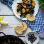Teller mit Muscheln und Brot auf blauem Geschirrtuch, Zitronenscheiben und Olivenschale daneben auf Holztisch.