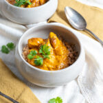 Schüssel mit cremigem Curry, Reis und Koriander auf weißer Tischdecke. Daneben liegt ein Metalllöffel auf einem gelben Tuch.