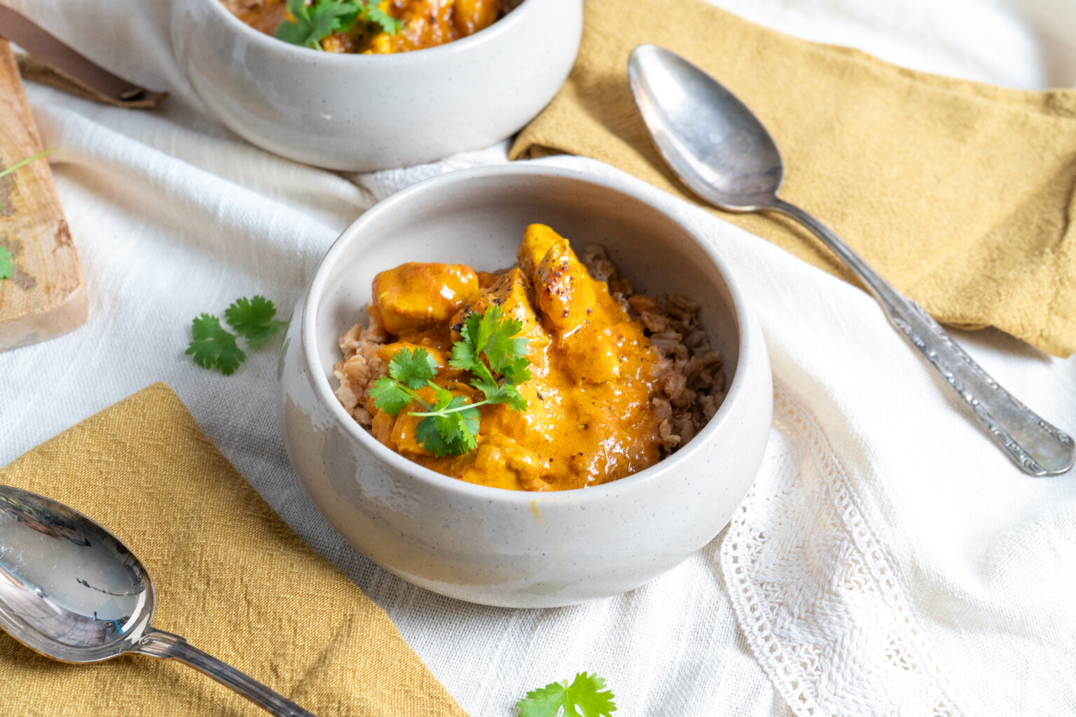 Schüssel mit cremigem Curry, Reis und Koriander auf weißer Tischdecke. Daneben liegt ein Metalllöffel auf einem gelben Tuch.