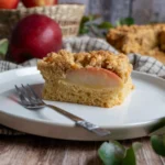 Stück Apfelkuchen mit Streuseln auf einem weißen Teller, Gabel daneben, im Hintergrund ein Korb mit Äpfeln.