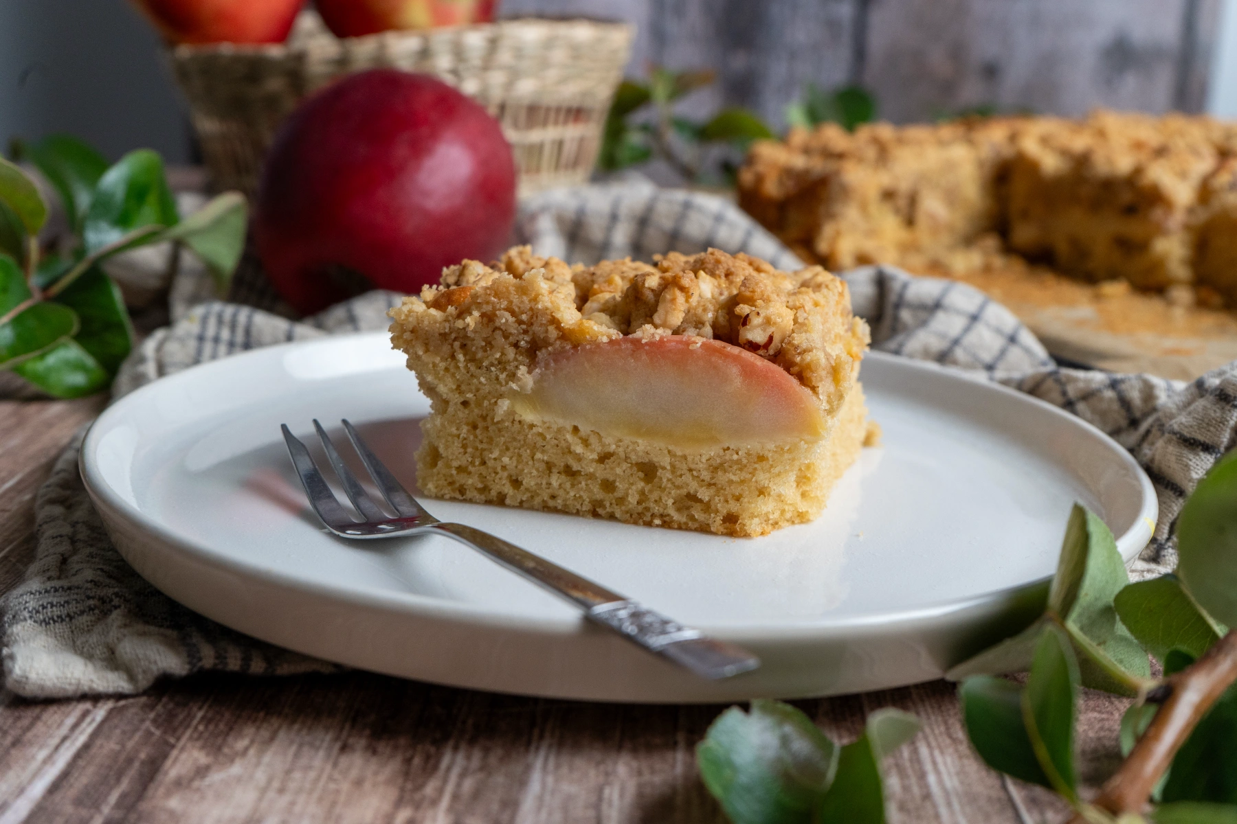 Ein Stück Apfelkuchen mit Streuseln auf einem weißen Teller, daneben eine Gabel und Äpfel im Hintergrund.