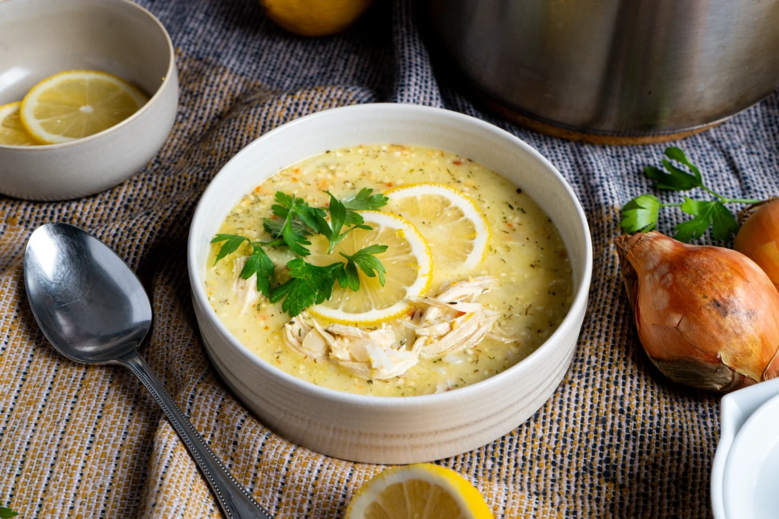 Cremige Suppe mit Zitronenscheiben und Kräutern, daneben Zwiebeln und ein Löffel, auf einem Stofftuch serviert.