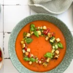 Tomatensuppe garniert mit Croutons, Basilikum und Gemüsewürfeln in einer Schüssel, auf einem Tisch mit Tomaten und Zitrone.