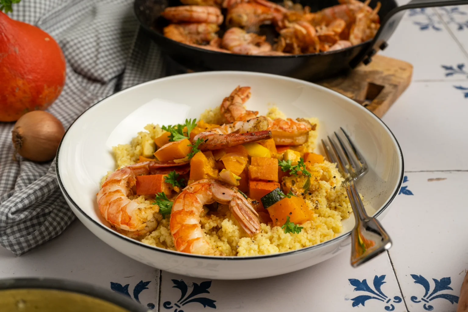 Ein Schüsselgericht mit Garnelen, Couscous und Kürbis, dekoriert mit Petersilie. Im Hintergrund eine Pfanne und Zwiebel.