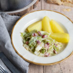 Teller mit Weißkohl, Speckstückchen und drei Salzkartoffeln, daneben graues Stofftuch und Besteck auf Holztisch.
