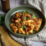 Grüner Teller mit Rigatoni in Fleischsauce, garniert mit Petersilie, auf Tisch mit gelbem Geschirrtuch und Gabel.