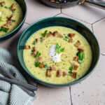 Zwei Schalen Zucchinisuppe mit Croutons, Kräutern und einem Klecks Sahne, daneben zwei Löffel auf einem Tuch.