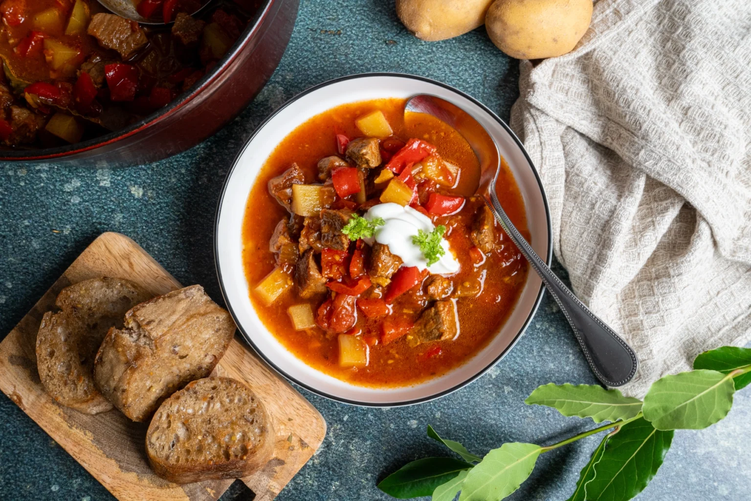 Eintopf mit Fleisch, Kartoffeln und Paprika in Schale, daneben Brot auf Schneidebrett, Serviette und Lorbeerblätter.