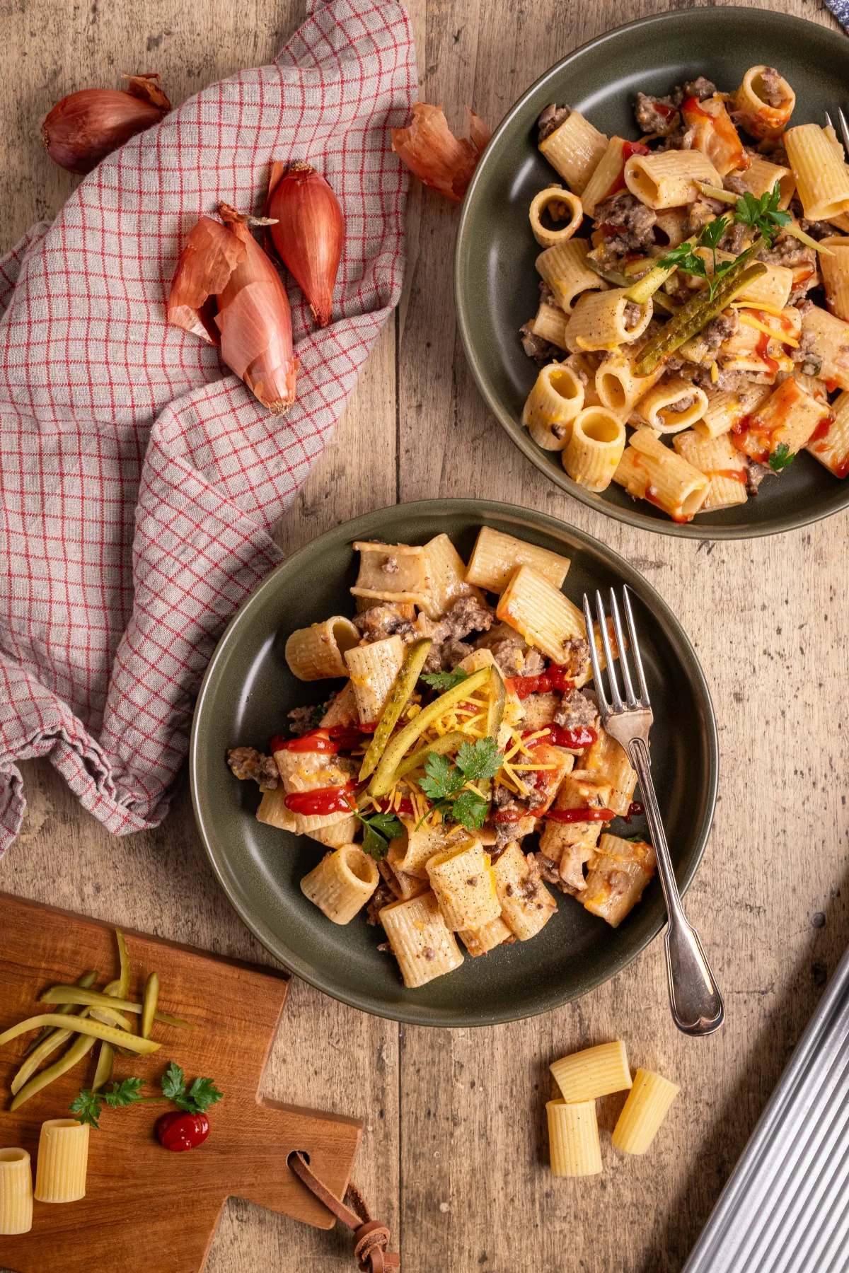 Zwei Teller mit Rigatoni, Gemüse und Kräutern auf Holztisch, flankiert von Schalotten und Küchenutensilien.