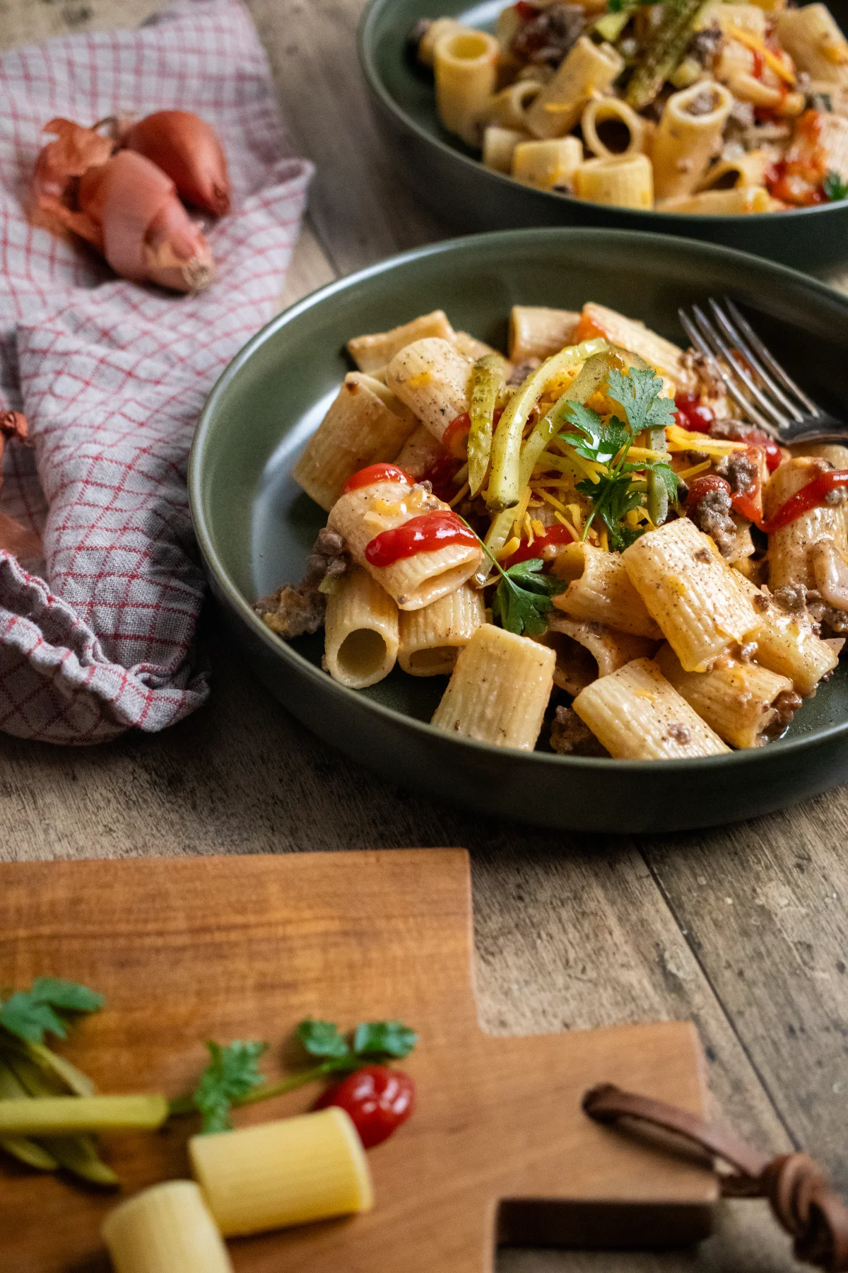 Grüner Teller mit Rigatoni, Hackfleisch, Paprika und Petersilie, auf Holztisch mit roten Zwiebeln und Geschirrtuch daneben.