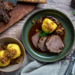 Gedeckter Tisch mit Braten, Soße, Kloß und Bröseln auf einem Teller, daneben ein Schneidebrett mit geschnittenem Fleisch.