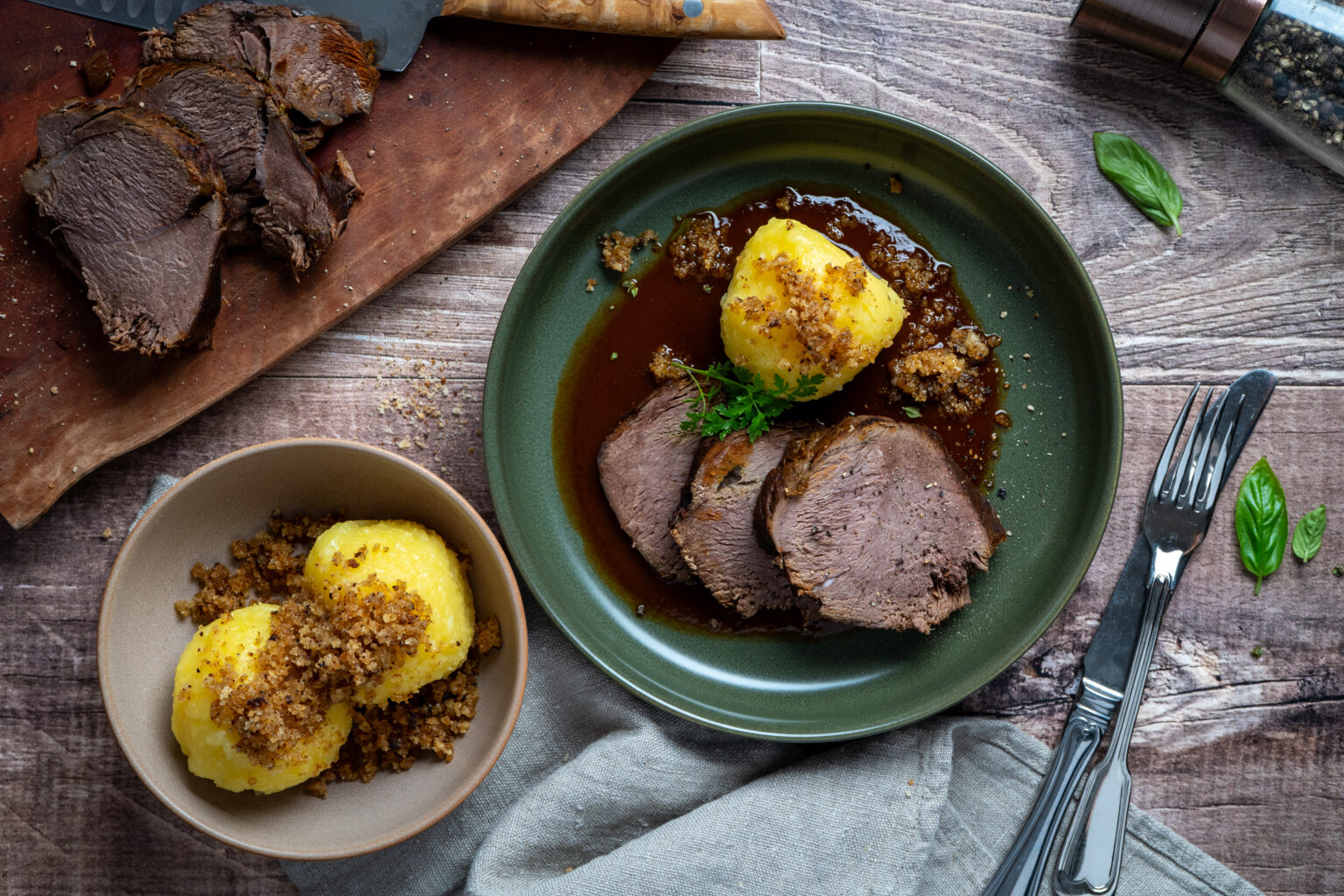 Gedeckter Tisch mit Braten, Soße, Kloß und Bröseln auf einem Teller, daneben ein Schneidebrett mit geschnittenem Fleisch.