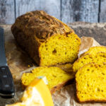 Aufgeschnittenes Kürbisbrot auf Backpapier, daneben ein Brotmesser und Kürbisstücke. Rustikaler Holzhintergrund.