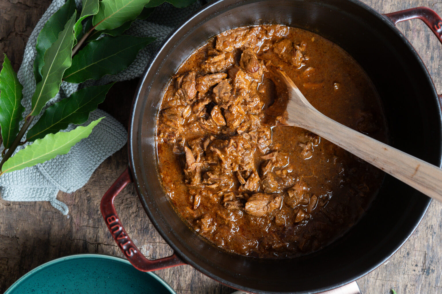 Gusseisener Topf mit Rindergulasch, Holzlöffel, grüne Blätter und graues Tuch daneben auf Holztisch.