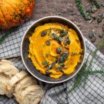 Kürbis-Dip von oben mit Kürbiskernen, daneben Baguette, Thymianzweige und ein Kürbis auf kariertem Tuch.