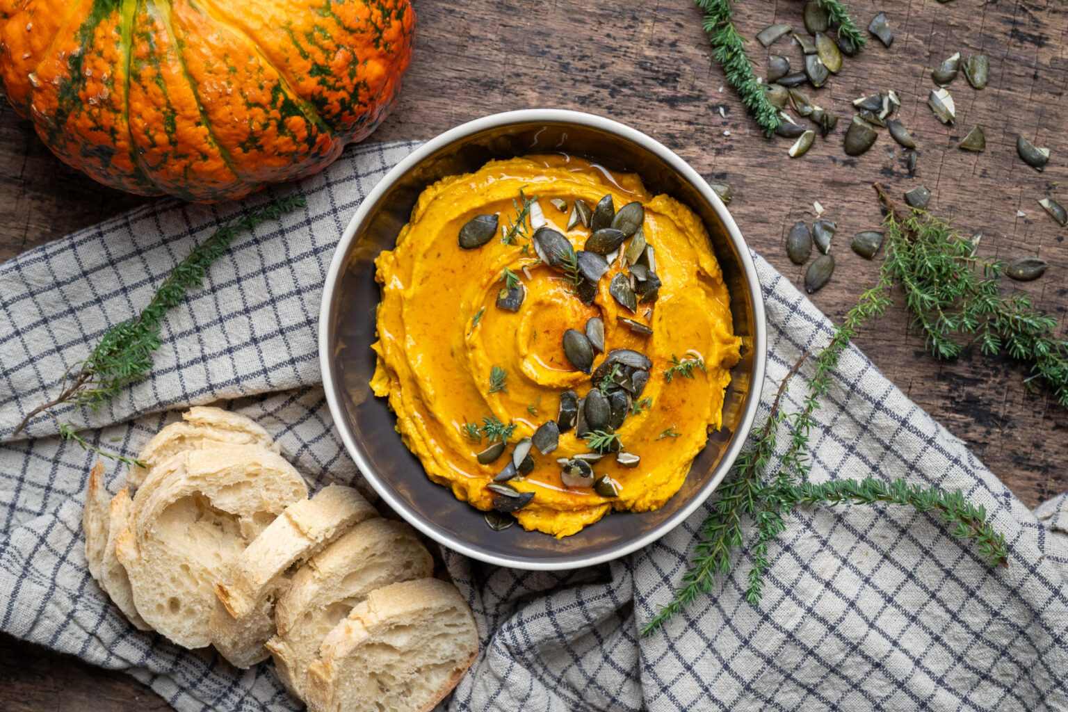 Kürbis-Dip von oben mit Kürbiskernen, daneben Baguette, Thymianzweige und ein Kürbis auf kariertem Tuch.