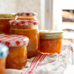 Mehrere Gläser mit selbstgemachter, orangefarbener Marmelade auf einem Küchentuch bei Tageslicht.