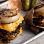 Zwei Cheeseburger mit gegrillten Zwiebeln, in Zeitungspapier gewickelt, auf einem Blech serviert.