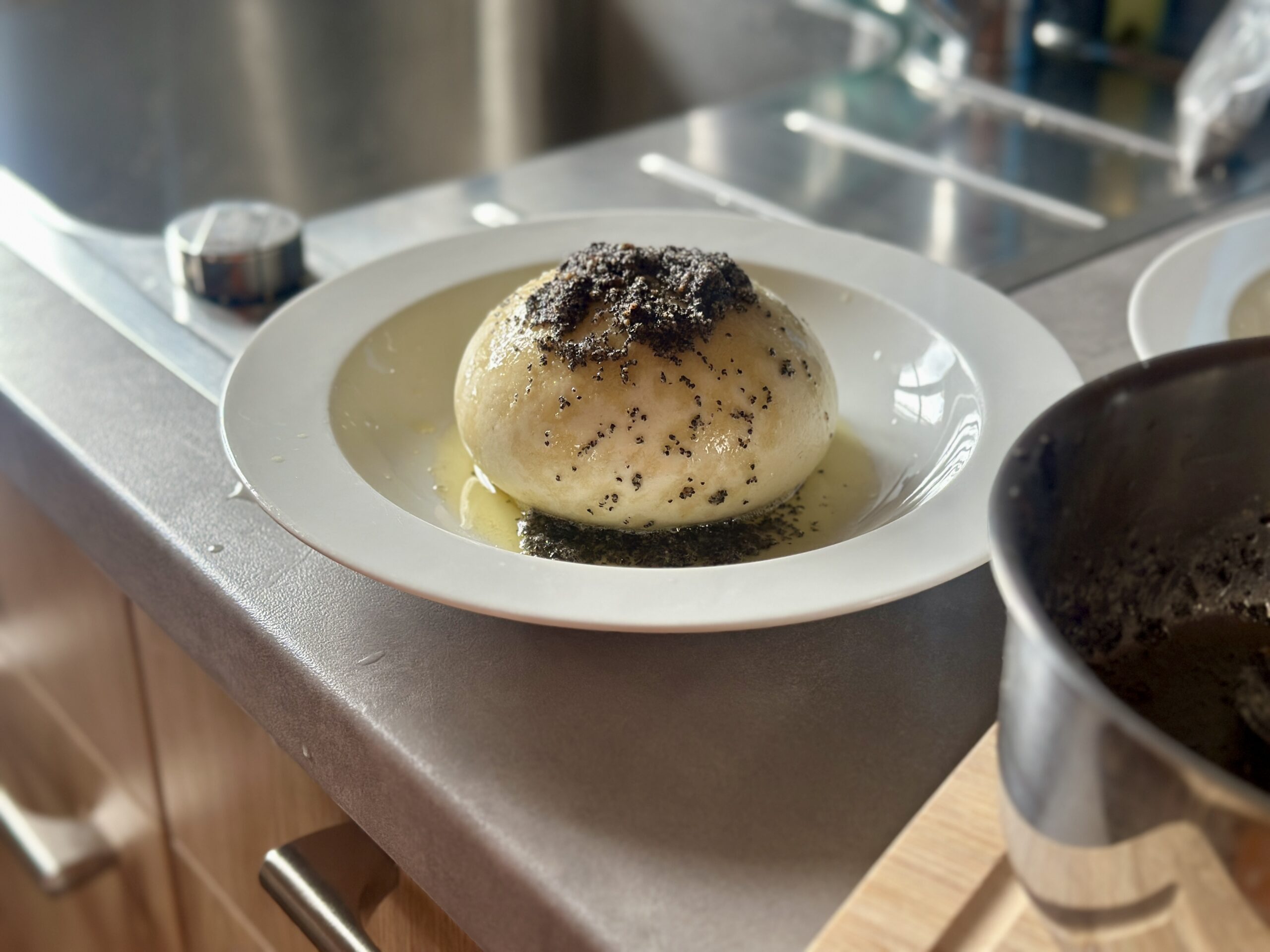Dampfnudel mit Mohn und Butter auf weißem Teller, daneben ein Topf auf einer Küchenarbeitsplatte.