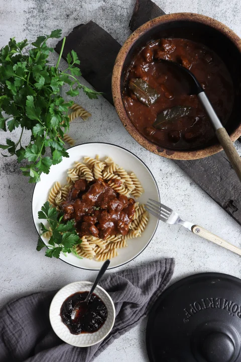 Teller mit Spiralnudeln und dunklem Eintopf, daneben ein Topf mit Eintopf, Petersilie oben links, Marmelade in kleinem Schälchen.