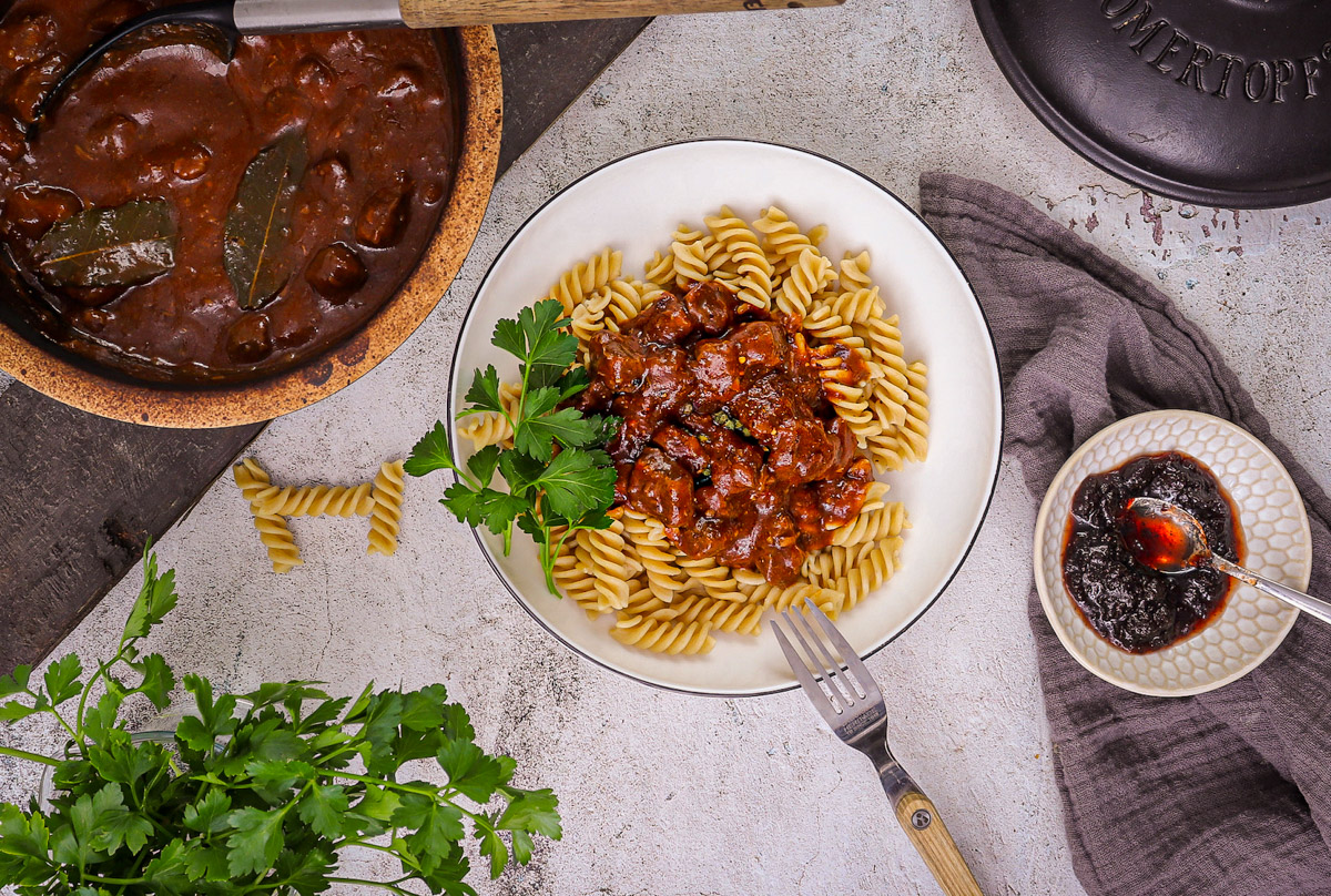 Gulasch-aus-dem-Römertopf-1 Teller mit Fusilli und Gulasch, garniert mit Petersilie, umgeben von Gulaschtopf und Schälchen mit dunkler Sauce.