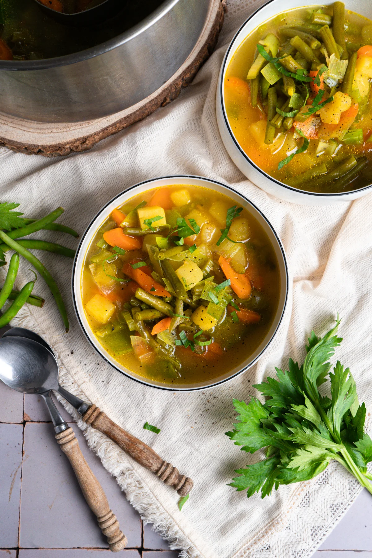 Zwei Schüsseln Gemüsesuppe mit Karotten, grünen Bohnen und Kartoffeln, daneben Petersilie und zwei Löffel auf Tuch.