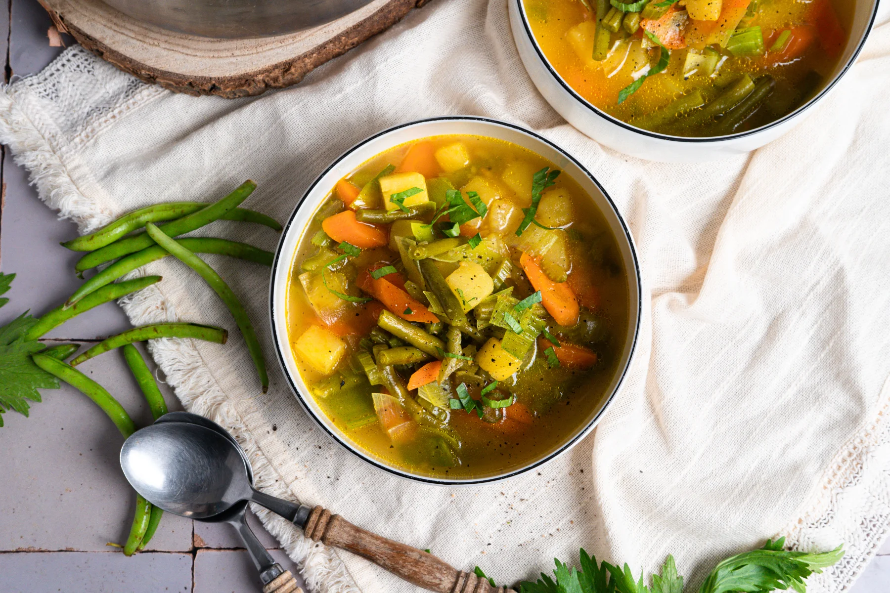 Eine Schüssel Gemüsesuppe mit Karotten, Bohnen und Kartoffeln auf einem Tuch; daneben Löffel und frische Bohnen.