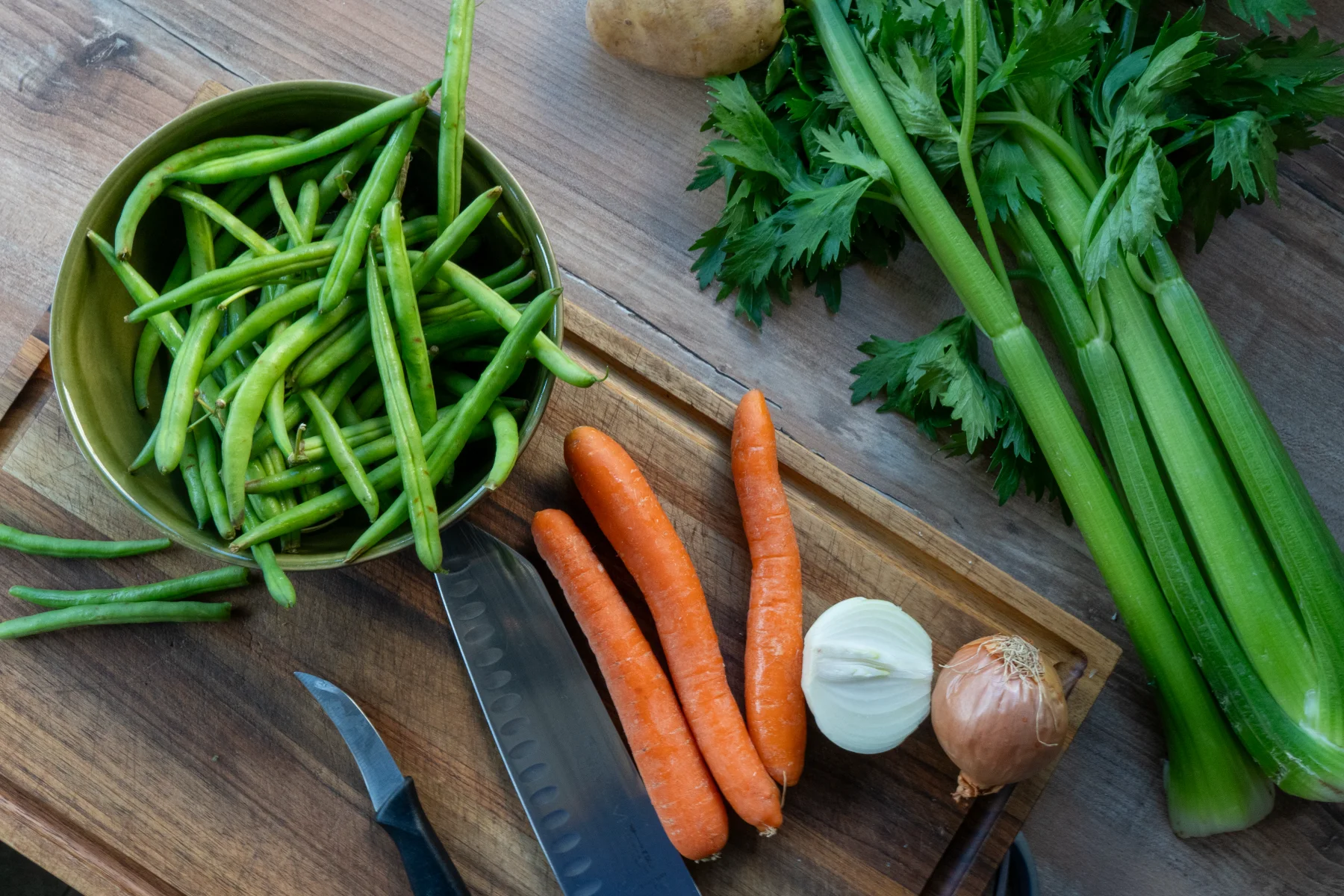 Frisches Gemüse: grüne Bohnen in Schüssel, Karotten, Zwiebeln, Sellerie und Messer auf Holzbrett.