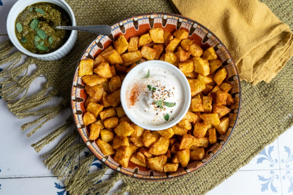 Würfelkartoffeln in einer Schale mit würzigem Dip, grünes Pesto und gelbe Serviette auf einem Tisch mit Stoffdecke.