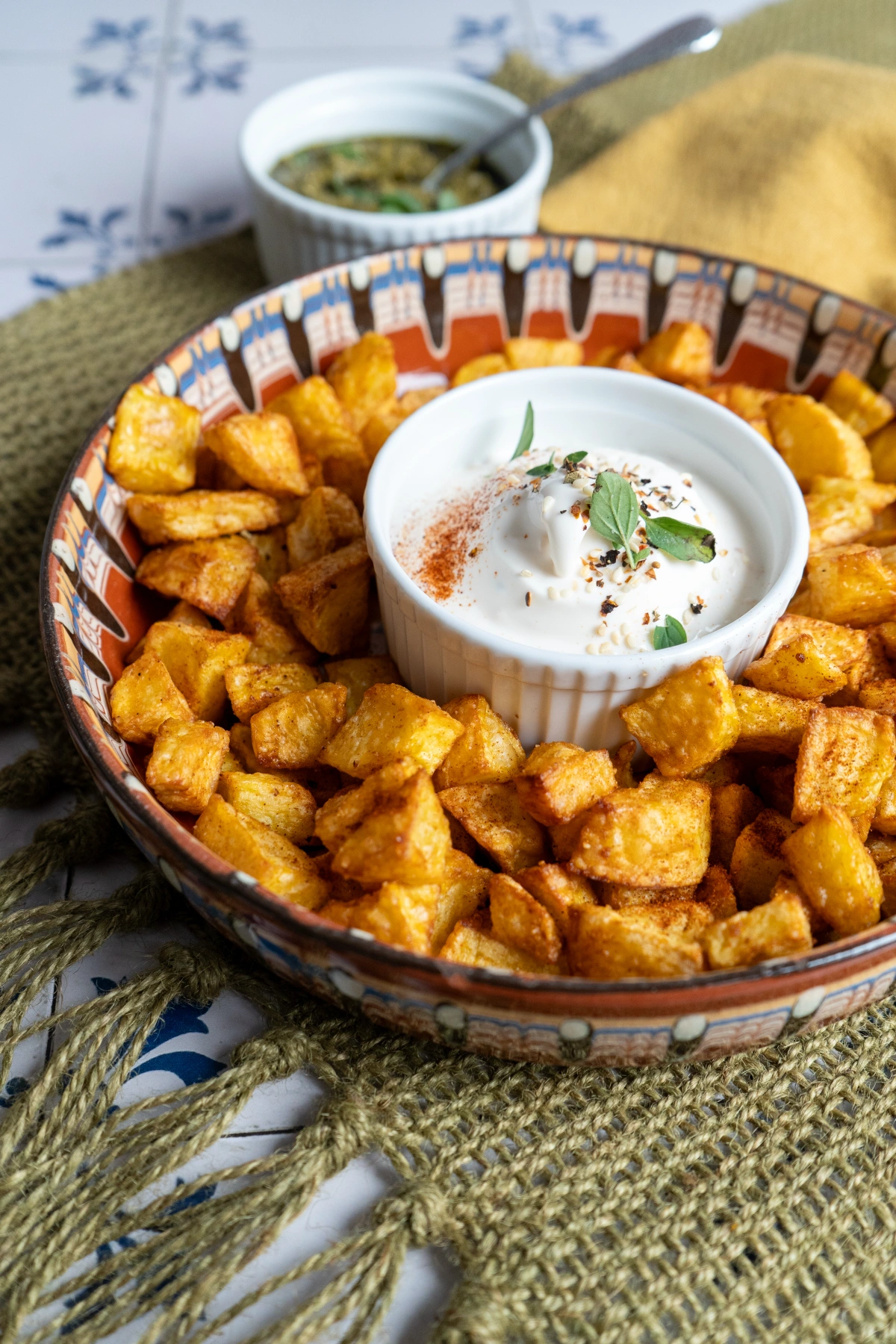 Knusprige Kartoffelwürfel in Schale mit Dip in der Mitte, dekoriert mit Kräutern, auf grünem Tuch serviert.