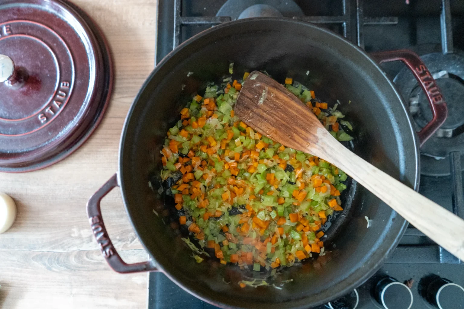 Karotten und Sellerie werden in einem Topf auf einem Herd angebraten, ein Holzlöffel liegt darin.