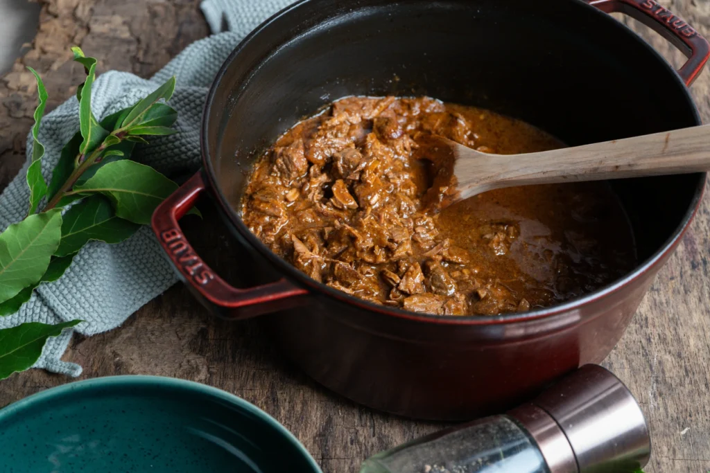Roter Topf mit Rindfleischgulasch auf Holzuntergrund, daneben Lorbeerblätter und ein blauer Teller.
